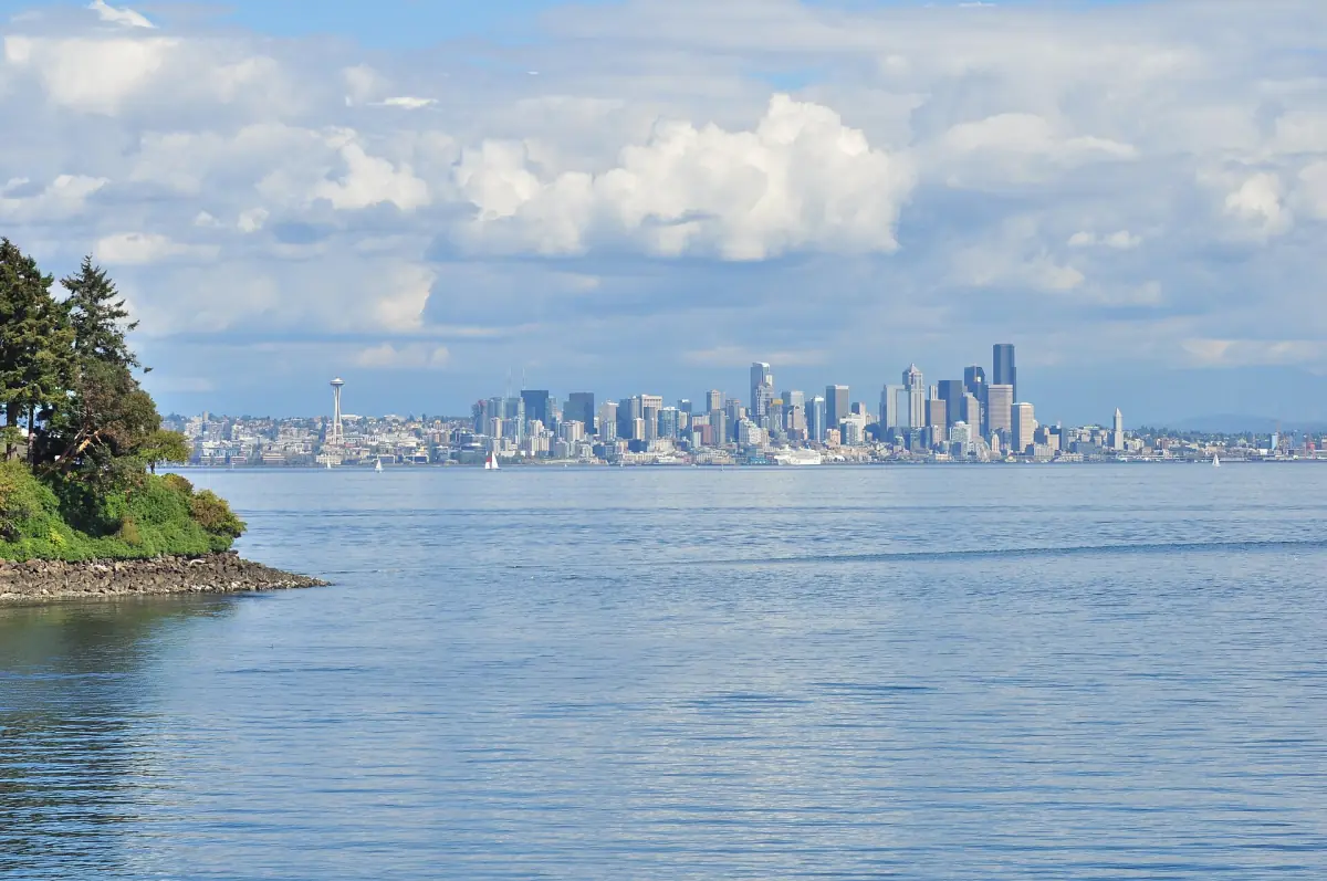 Distant Seattle skyline over calm water with a wooded point in the foreground